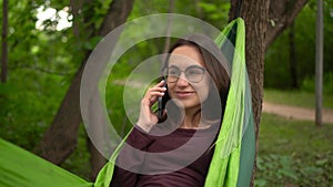 Young woman in glasses talking on the phone while lying in a hammock. A girl with glasses is resting in a hammock in