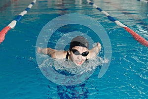 Young woman in goggles and cap swimming butterfly stroke style in the blue water indoor race pool