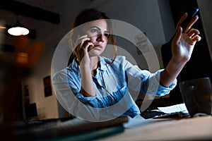 Young woman graphic designer using pc computer talking by mobile phone working at night in office.