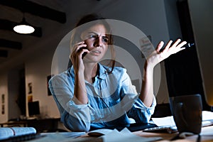 Young woman graphic designer using pc computer talking by mobile phone working at night in office.