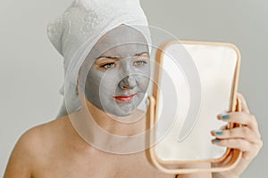 Young woman with gray clay cosmetic mask on her face looks in mirror, her hair and body wrapped in towel.