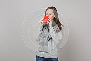 Young woman in gray sweater, scarf drinking coffee or tea from red cup isolated on grey background in studio. Healthy