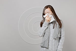 Young woman in gray sweater, scarf having runny nose, blowing nose to napkin isolated on grey wall background. Healthy