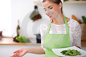 Young woman in green apron is cooking in a kitchen. Housewife is offering fresh salad.