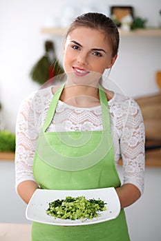 Young woman in green apron is cooking in a kitchen. Housewife is offering fresh salad.