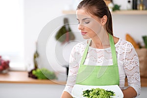 Young woman in green apron is cooking in a kitchen. Housewife is offering fresh salad.