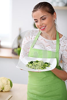 Young woman in green apron is cooking in a kitchen. Housewife is offering fresh salad.