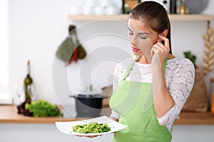 Young woman in green apron is cooking in a kitchen. Housewife is offering fresh salad.