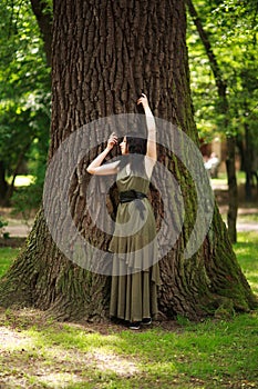 Young woman in green dress meditatively relaxes near large tree in forest park, concept of purity of nature and unity with