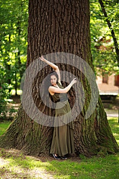 Young woman in green dress meditatively relaxes near large tree in forest park, concept of purity of nature and unity with