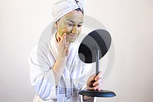 Young woman with a green mask on her face in a bathrobe in front of a mirror.