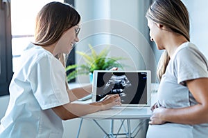 Young woman gynecologist doctor showing to pregnant woman ultrasound scan baby with digital tablet in medical consultation