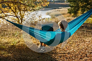 Young woman in hammok using laptop working outdoor facing lake on a sunset