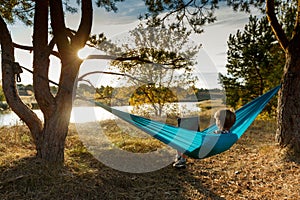 Young woman in hammok using laptop working outdoor facing lake on a sunset