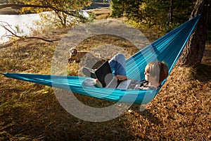 Young woman in hammok using laptop working outdoor facing lake on a sunset together with her home pet dog