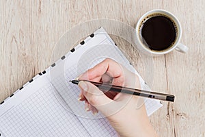 Young woman hand hold opened notebook pages with black pen next to cup of coffee on  wooden table. Top view