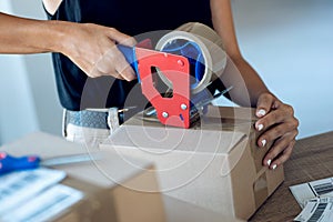 Young woman hands holding packing machine and sealing cardboard boxes with duct tape to delivery products ordered online to