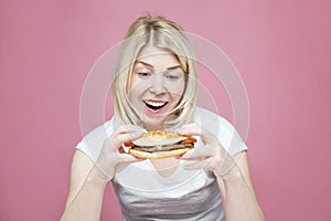 A young woman happily eats a hamburger. Beautiful blonde in a white shirt. Hunger, diets and junk delicious food. Pink background