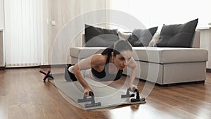 Young woman has workout at home during quarantine. Strong powerful girl dong push ups with equipment. Confident sporty