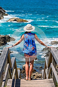 A young woman in a hat and a blue T-shirt stands on wooden steps and looks at the blue sea with rocks, seaside landscape