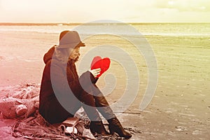 A young woman in a hat and coat sits on the shore of the bay on a fishing net with a plush heart in her hands, Bright toning, copy