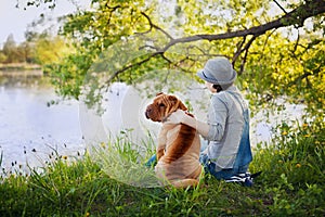 Young woman in a hat with dog Shar Pei sitting in the field and looking to the river in golden sunset light