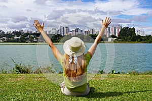 Young woman with hat raising arms sitting on grass in Barigui Park, Curitiba, Brazil photo