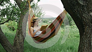 Young woman with hat resting in comfortable hammock at green garden. Cute charming brunette female reading book lying in hammock o