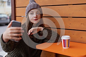 Young woman in hat and sweater sitting on the outdoor cafe, using smartphone, making selfie and drinking coffee