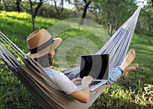 Young woman in a hat using a laptop while lying in a hammock in a summer garden. Summer lifestyle