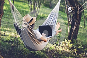 Young woman in a hat using a laptop while lying in a hammock in a summer garden. Summer lifestyle concept, working online