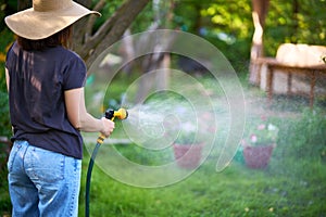 Young woman in hat watering flowers and plants in garden with hose in sunny blooming backyard