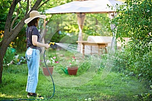 Young woman in hat watering flowers and plants in garden with hose in sunny blooming backyard