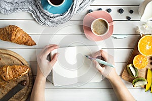 Young woman have a breakfast with fresh croissants, coffee and fruits and her hands drawing or writing with ink pen