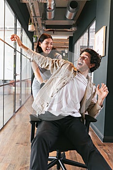 Young woman having fun pushing man on chair along walkway in open plan office