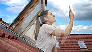 Young woman having issues with lost signal of a cellphone or smartphone looking out of the open attic window and trying