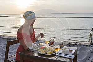 Young woman having romantic dinner in hotel restaurant during sunset near sea waves on the tropical beach, close up