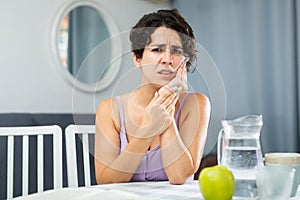 Young woman having toothache and touching cheek, sitting at table