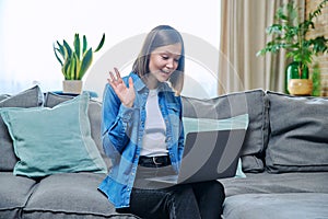 Young woman having video call sitting on sofa at home waving, using laptop