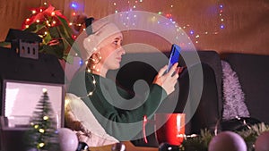 Young woman in headphones and hat sitting on the sofa in Christmas decorated living room using mobile phone.