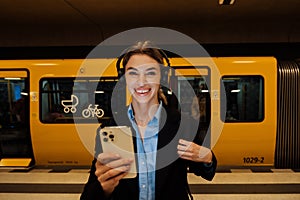 Young woman in headphones holding mobile phone while waiting for train at subway station