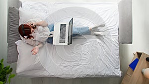 young woman with headset and smartphone working at laptop while lying on bed in bedroom, top view