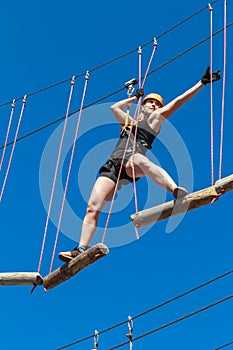 Young woman in a helmet with insurance in an adventure climbing park walks by a rope simulator against a blue sky