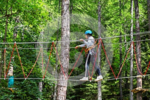 Young woman in helmet walking on suspended wires in forest adventure park