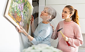 Young woman helping elderly mother to hang painting on wall