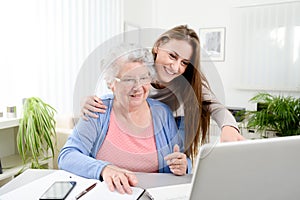 Young woman helping an old senior woman doing paperwork and administrative procedures with laptop computer at home