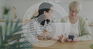 Young woman helping senior pensioner with paperwork and paying bills talking in kitchen at home