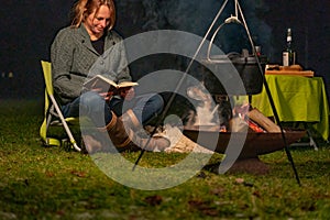 Young woman and her Australian Shepherd outside by a campfire. Reading a book at dusk. Bread, cheese and wine on the