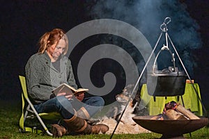 Young woman and her Australian Shepherd outside by a campfire. Reading a book at dusk. Bread, cheese and wine on the