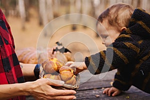 Young woman and her baby son in autumn park, boy playing with helloween pumpkin and eating pumpkin bun.